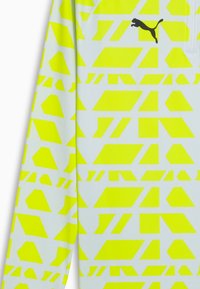 Sporttröja i lättviktsmaterial, med ett neongul geometrisk mönster på vit bakgrund, och en svart Puma-logotyp.