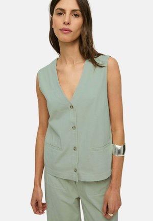 Mujer con top sin mangas abotonado de color verde claro y pantalones a juego, con brazalete de plata en la muñeca izquierda y anillo en la mano derecha.