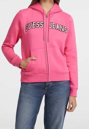 Kobieta ubrana w różową bluzę na suwak z napisem „GUESS JEANS”, z jedną ręką w przedniej kieszeni, w połączeniu z niebieskimi dżinsami.