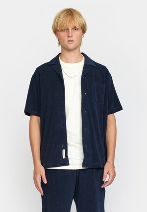 Marineblauw overhemd met korte mouwen, gemaakt van zachte stof, met een kraag, knopen aan de voorkant en een borstzak, gedragen over een wit t-shirt.