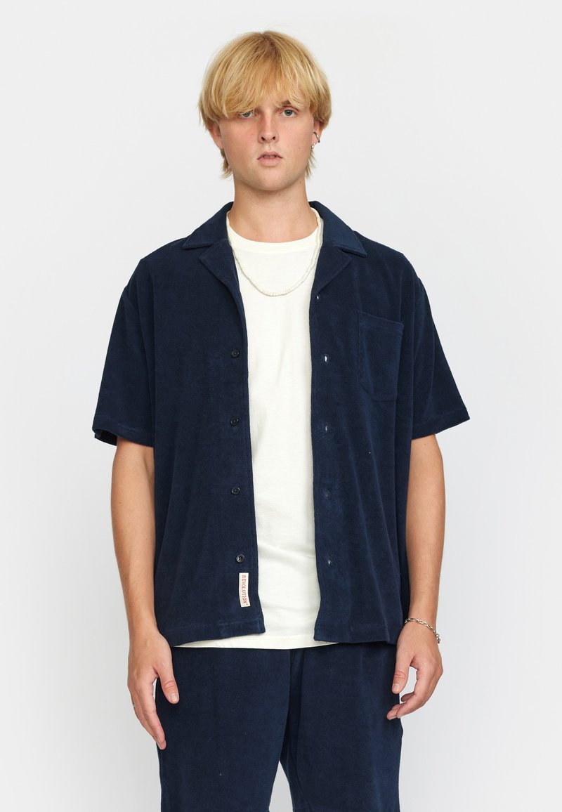 Marineblauw overhemd met korte mouwen, gemaakt van zachte stof, met een kraag, knopen aan de voorkant en een borstzak, gedragen over een wit t-shirt.