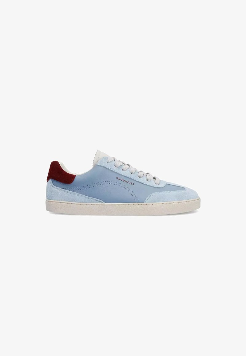 Baskets basses bleu clair avec lacets blancs, semelle beige, accents en daim et pièce de talon rouge foncé, étiquetées « GROUNDIES » sur le côté.