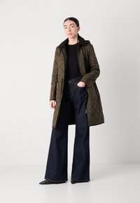 Olijfgroene gevoerde lange jas met capuchon, zwarte voering en ritssluiting. Gecombineerd met hoge taille donkere spijkerflare jeans en zwarte puntige schoenen.