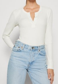 Vit ribbad långärmad topp med en liten knappslå, tillsammans med högmidjade ljusblå jeans med knappar och fickdetaljer.