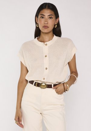 Frau trägt beiges Kurzarm-Strick-Button-up-Top, cremefarbene hoch taillierte Hose, brauner Gürtel mit großer goldener Schnalle und goldener Schmuck.