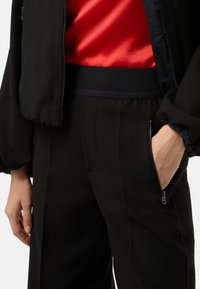Czarna kurtka typu bomber i pasujące spodnie z fakturowanym paskiem w talii. Spodnie mają boczne kieszenie i gładkie wykończenie materiału. Pod spodem widoczna czerwona satynowa koszula.