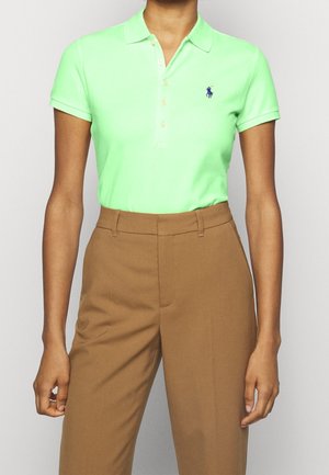 Femme portant un polo vert clair à manches courtes, rentré dans un pantalon marron taille haute.