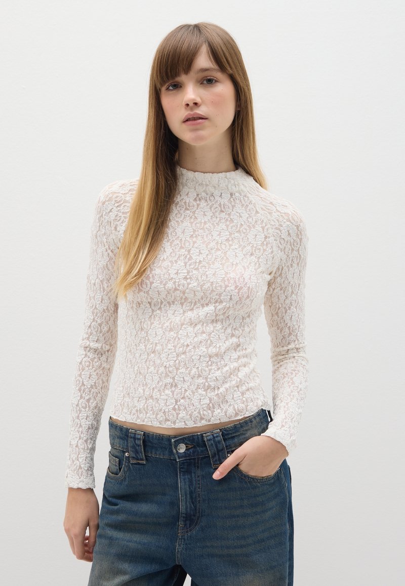 Top bianco in pizzo a maniche lunghe con collo alto, caratterizzato da motivi floreali e una silhouette aderente, abbinato a jeans blu scuro a vita alta.