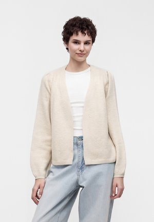 Jeune femme aux cheveux courts et bouclés, portant un haut blanc, un cardigan beige et un jean bleu clair, debout devant un fond uni.