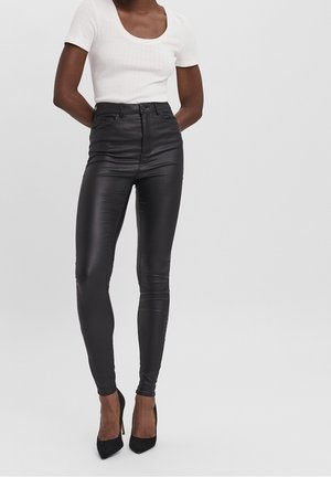 Schwarze, hoch taillierte Skinny-Hosen aus Kunstleder mit glänzender Oberfläche, kombiniert mit einem weißen Kurzarmoberteil mit Rundhalsausschnitt und schwarzen High Heels.