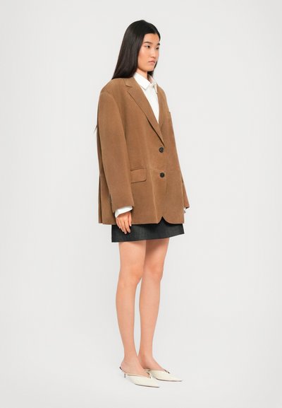Donna in piedi di lato che indossa un blazer marrone oversize, una camicia bianca, una gonna nera e delle mule bianche con tacco a punta su uno sfondo neutro.