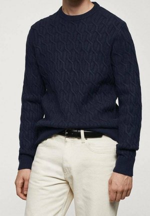 Marineblau gestrickter Pullover mit strukturiertem Kabelmuster. Rundhals, lange Ärmel und gerippte Bündchen. Kombiniert mit hellfarbigen Hosen und einem schwarzen Gürtel.