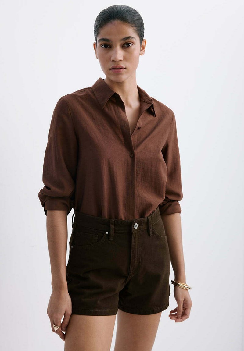 Femme aux cheveux foncés en chignon portant une chemise marron à manches longues boutonnée et un short marron foncé taille haute, debout devant un fond uni.