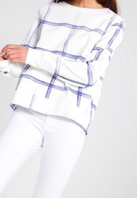 Sudadera blanca con diseño de cuadros azules, de manga larga y corte holgado. Presenta un cuello redondo y aberturas laterales en el dobladillo.