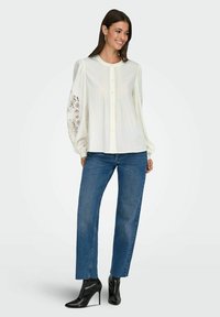 Biała bluzka z guzikami, z teksturowanymi koronkowymi rękawami, w zestawie z ciemnoniebieskimi prostymi dżinsami i czarnymi botkami na wysokim obcasie.