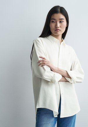 Młoda kobieta o długich ciemnych włosach, ubrana w luźną białą koszulę na guziki z podwiniętymi rękawami i niebieskie dżinsy, stojąca na jednolitym tle.