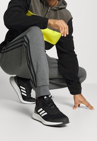 Sort og grå fleece hoodie med gul stribe, grå sweatpants med sorte sidestiber og sorte atletiske sko med hvide detaljer.