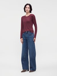 Femme aux cheveux courts et foncés portant un haut bordeaux à manches longues, un jean bleu à jambes larges et des chaussures pointues marron, debout devant un fond uni.