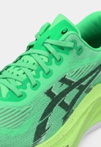 Neon zöld futócipő könnyű hálós felsőrésszel, texturált ráhajtásokkal, fekete logóval és párnázott talppal, mely pöttyös mintázatot tartalmaz.