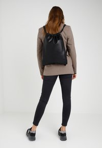 Femme portant un blazer à carreaux, un jean skinny noir, des baskets noires et un sac à dos noir à cordon, debout contre un fond blanc uni.