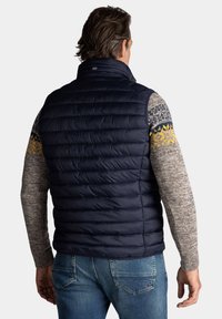 Marineblauwe gewatteerde vest met een gestikt ontwerp, gecombineerd met een gestructureerde, gebreide trui met gele en blauwe accenten.