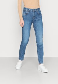 Jeans de ganga azuis com um corte justo, design de cinco bolsos, desbotamento leve e cintura média, combinados com ténis brancos.