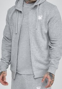 Sudadera gris con cremallera hecha de un tejido suave, que presenta una capucha con cordones, puños acanalados y un pequeño logo blanco en el pecho.