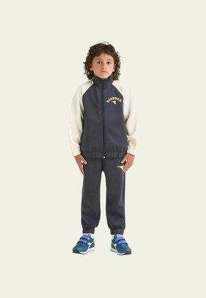 Bambino che indossa una tuta blu navy con maniche bianche e logo giallo. Pantaloni slim-fit con polsini elastici; le scarpe presentano dettagli blu e cinturini in Velcro.