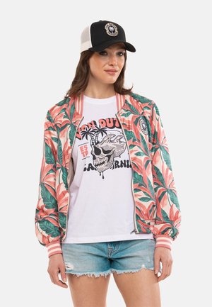 Femme portant une casquette noire et blanche, une veste bomber fleurie rose et verte, un t-shirt blanc avec un crâne graphique et un short en jean effiloché.