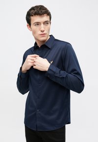 Donkerblauw overhemd met lange mouwen, een button-down kraag, subtiele structuur en een klein goudkleurig logo op de linkerborst. Zwarte broek zichtbaar.