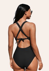 Il costume da bagno intero nero presenta un design con schiena incrociata e dettaglio a fiocco, gambe alte e una texture del tessuto liscia.