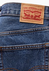 Gros plan sur la poche arrière d'un jean en denim bleu avec des coutures orange et un patch en cuir marron Levi Strauss représentant deux chevaux et du texte.