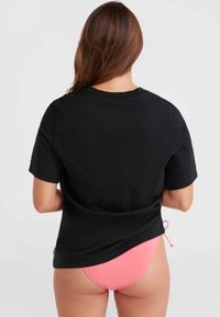 Sort bomulds t-shirt med korte ærmer, der har en afslappet pasform. En pink bikini underdel er synlig under skjorten.
