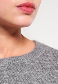 Grauer gestrickter Pullover mit rundem Ausschnitt. Nahaufnahme von Lippen und Kinn, die die weiche Textur des Stoffes und die neutrale Farbe hervorhebt.