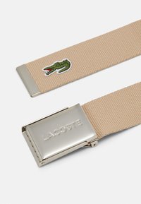 Cinturón de lona beige con un logo de cocodrilo bordado en verde y una hebilla de metal plateada que lleva la palabra "LACOSTE". Se aprecia un detalle de tela texturizada.