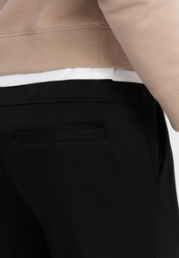 Schwarze Jogginghosen mit einer Gesäßtasche, kombiniert mit einem beigen Sweatshirtsaum, der ein darunter liegendes weißes Shirt zeigt. Weiche Stofftextur.