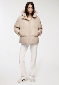 Beżowa kurtka puchowa z wysokim kołnierzem i zapinana na zamek, z dwoma dużymi kieszeniami oraz gładką, błyszczącą teksturą. W połączeniu z białymi spodniami.