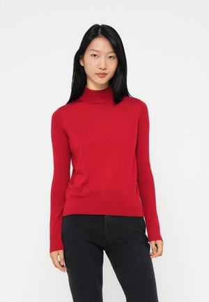 Woman with long black hair wearing a red turtleneck sweater and black pants, standing against a white background.