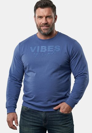 Mężczyzna z krótkimi brązowymi włosami i brodą, ubrany w niebieską bluzę z tekstem "Vibes of Cph" i niebieskie jeansy, stoi na jednolitym tle.