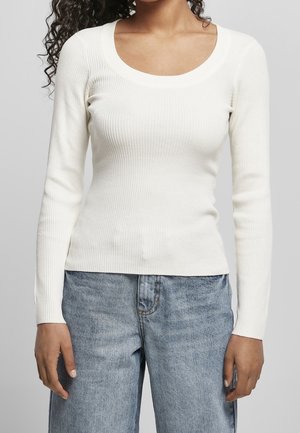 Vrouw die een wit geribd langemouwen topje en lichtblauwe jeans met hoge taille draagt, staand tegen een effen grijze achtergrond.