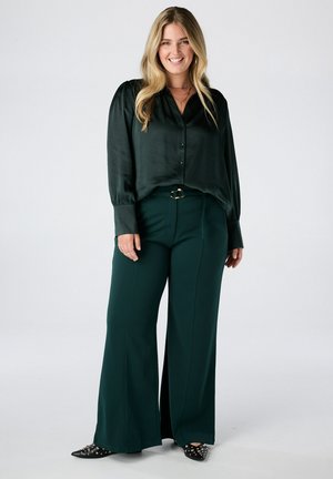 Lachende vrouw met lang blond haar staat, gekleed in een donkergroene satijnen blouse, wijde bijpassende broek en zwarte schoenen met studs.