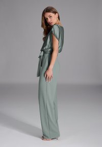 Jumpsuit verde in satin con vestibilità ampia, maniche corte, vita con cintura e pantaloni a gamba larga. Texture liscia e design minimalista.