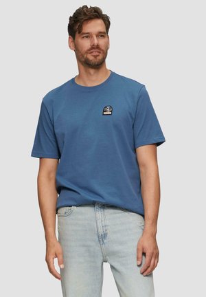 T-shirt en coton bleu avec un col rond et des manches courtes, featuring un petit patch brodé sur la poitrine. Porté avec un jean en denim clair.
