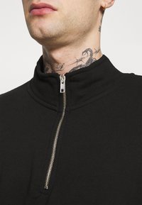 Zwarte sweatshirt met rits en hoge kraag. Voorzien van een zilveren rits, een gladde textuur en minimale naden. Het lichaam lijkt vanaf de nek naar beneden aansluitend.