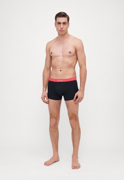 Boxer-briefs noirs avec une ceinture rouge vif, en tissu lisse et ajusté, présentés sur un modèle masculin sans chemise.