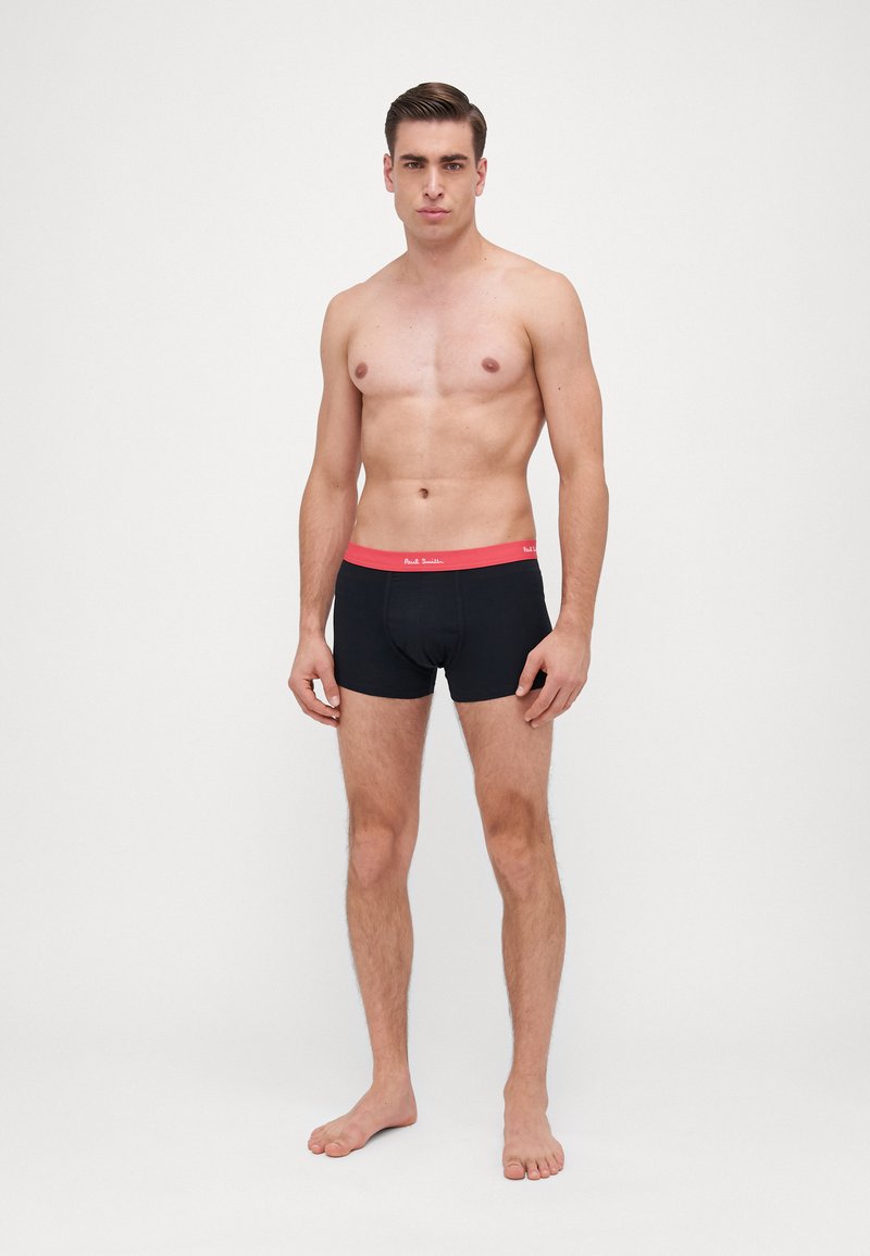 Boxer-briefs noirs avec une ceinture rouge vif, en tissu lisse et ajusté, présentés sur un modèle masculin sans chemise.