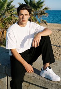 Junger Mann in weißem T-Shirt, schwarzen Hosen und weißen Turnschuhen, der auf einer Betonmauer am Strand mit Palmen und Meer sitzt.