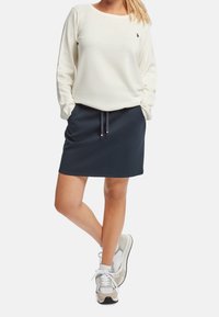 Pull crème à manches longues, associé à une jupe navy à cordon. Chaussures : baskets gris clair avec des accents foncés.