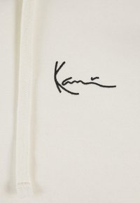 La tela de la sudadera blanca presenta una textura suave, con la firma bordada en negro "Kani" en el frente. Incluye cordones en el cuello.