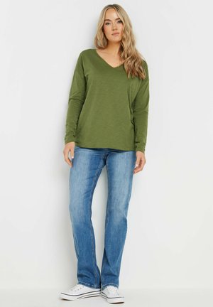 Haut vert à manches longues et col en V associé à un jean droit bleu et des baskets blanches ; texture douce avec une coupe décontractée.